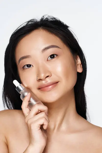 Young asian woman showcases a beautiful smile while holding a skincare serum close to her face. — Stock Photo