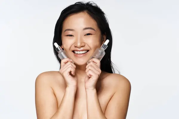 Smiling young woman holds skincare serums, radiating beauty and confidence in a bright studio. — Stock Photo