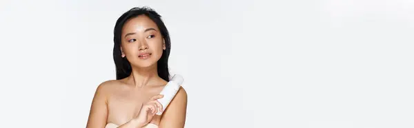 Una giovane donna asiatica si alza con fiducia, tenendo in mano un prodotto di bellezza e mostrando la sua pelle radiosa. — Foto stock