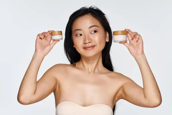 Cheery young Asian woman holds skincare jars, radiating beauty and confidence in a bright space. — Stock Photo