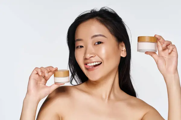 Smiling young Asian woman promotes natural beauty with skincare jars in a bright setting. — Stock Photo