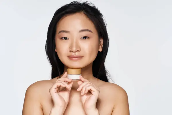 A young beautiful Asian woman smiles while holding a skincare jar, highlighting her natural beauty — Stock Photo