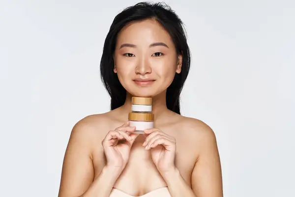 Brightly lit environment features a young asian woman presenting skincare jars with joy and poise. — Stock Photo