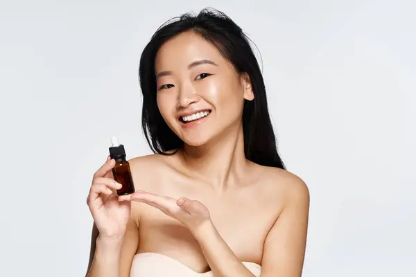 Une jeune femme asiatique joyeuse présente un sérum de soin avec un sourire radieux. — Photo de stock