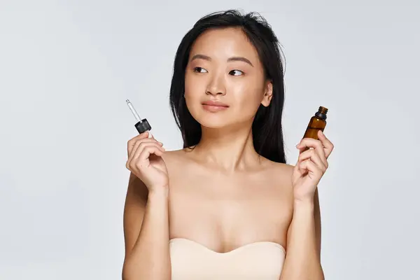 Young Asian woman smiles while holding two beauty serums, highlighting her skincare routine. — Stock Photo