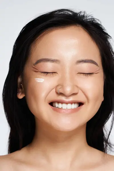 A young Asian woman smiles with closed eyes, radiating joy while applying skincare to her cheeks. — Stock Photo
