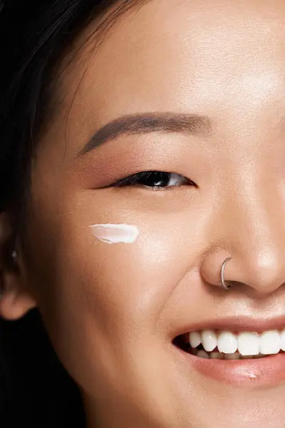 Young asian woman applies cream on her cheek while showcasing her bright and joyful smile. — Stock Photo