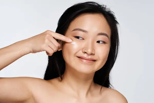 A cheerful young Asian woman applies moisturizer to her cheek, glowing with confidence and beauty. — Stock Photo