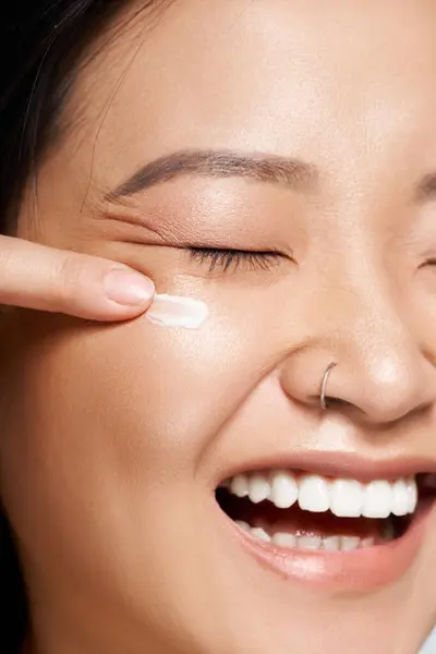 Sourire lumineux améliore la beauté naturelle comme une jeune femme asiatique applique de la crème sur sa joue. — Photo de stock