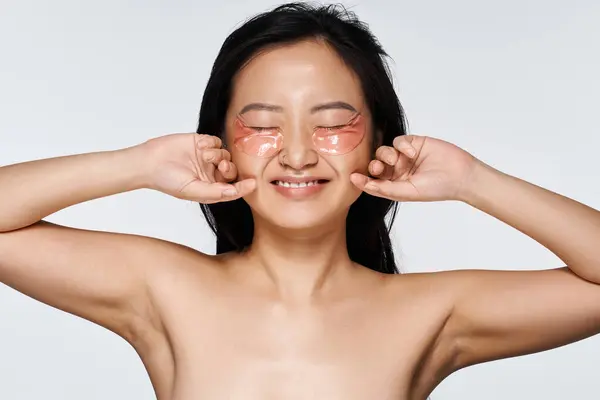 Bright smile from a young Asian woman as she applies eye patches, embracing self care and joy. — Stock Photo