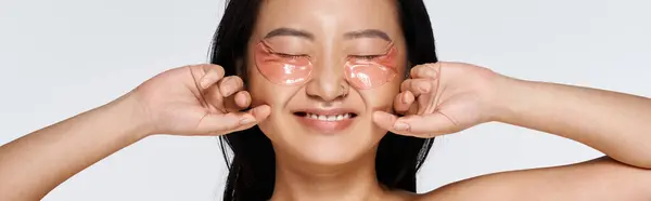 A young Asian woman happily applies under eye masks, enjoying a moment of self care. — Stock Photo