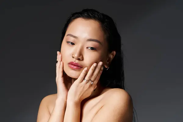 Young Asian woman showcases her glow, gently touching her face in soft light. — Stock Photo