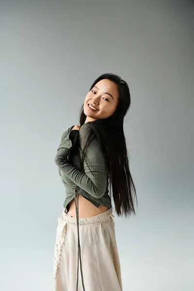 Una joven asiática sonríe mientras muestra juguetonamente su elegante atuendo en un sereno ambiente de estudio. - foto de stock