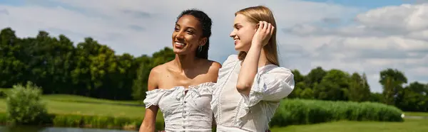 Duas jovens mulheres compartilham risos e carinho enquanto caminham perto de um lago sereno em uma tarde ensolarada. — Fotografia de Stock