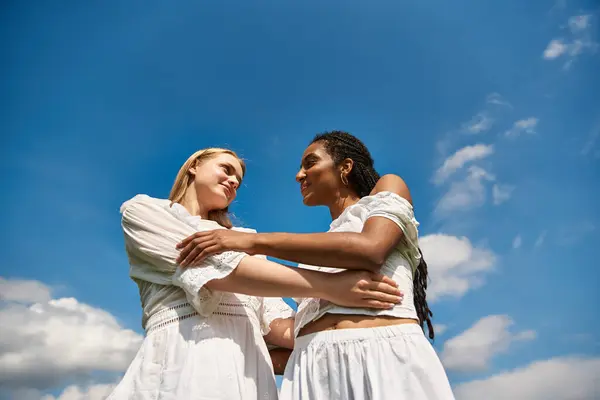 Duas jovens mulheres abraçam carinhosamente em um ambiente verde exuberante enquanto as nuvens vagam preguiçosamente por cima. — Fotografia de Stock