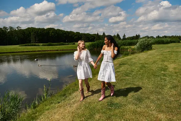 Two young women walk hand in hand along a serene lakeside, sharing laughter and love amidst nature. — Stock Photo