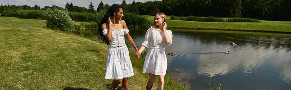 Two young women walk hand in hand by a tranquil pond, radiating love and warmth in nature. — Stock Photo
