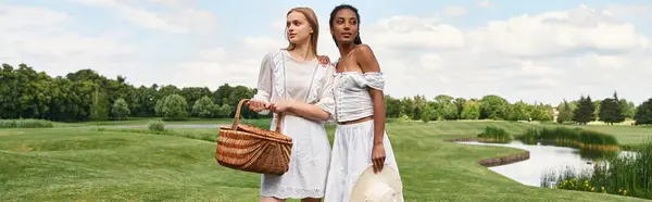 Two beautiful young women stand together, sharing smiles while holding a picnic basket in the park. — Stock Photo