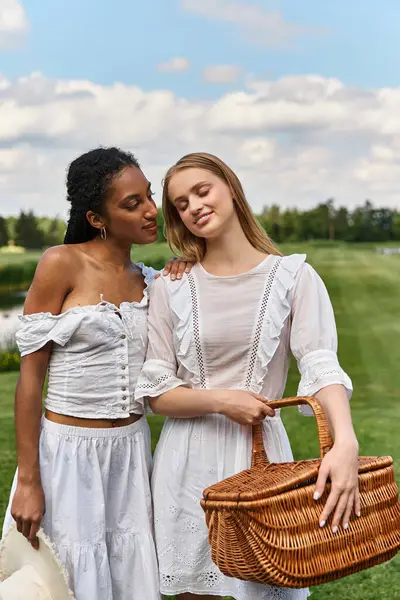 Two beautiful young women embrace lovingly while holding a picnic basket in a vibrant green field. — Stock Photo