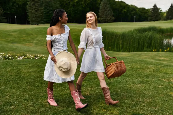 Two young women happily walk hand in hand in a grassy park, wearing summer dresses. — Stock Photo