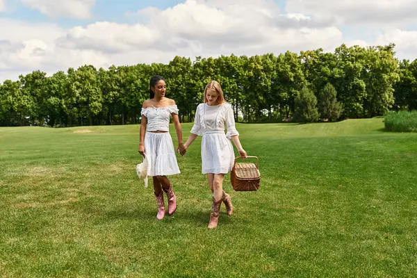 Two young women in white dresses enjoy a romantic stroll through a lush green park, holding hands. — Stock Photo