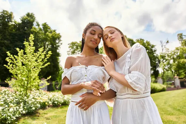 Two young women dressed in white share a tender moment in a lush, vibrant garden under the sun — Stock Photo