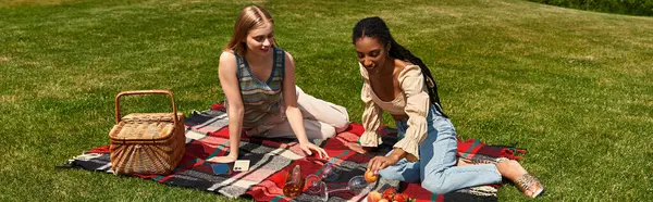 Two joyful young women sit on a picnic blanket, sharing fruits and smiles under the warm sun — Stock Photo
