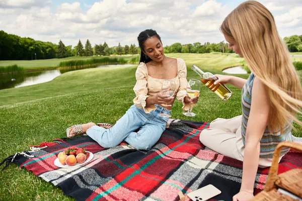 Duas jovens mulheres compartilham risos e coquetéis enquanto fazem piqueniques em um cobertor em um parque cênico. — Fotografia de Stock