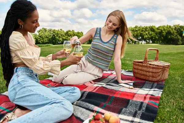 Duas jovens mulheres desfrutam de um dia ensolarado no parque, compartilhando risos e bebidas em um cobertor colorido. — Fotografia de Stock