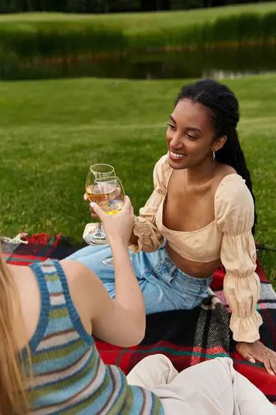 Two beautiful women share laughter and drinks while relaxing on a picnic blanket in a scenic park — Stock Photo