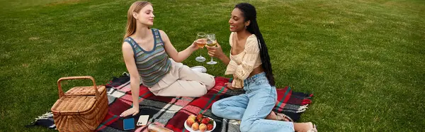 Two women share a joyful toast with glasses while sitting on a picnic blanket in a lush green park. — Stock Photo