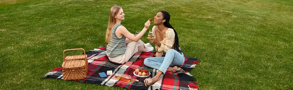 Dos mujeres jóvenes disfrutan de un acogedor picnic juntas, alimentándose mutuamente con alegría y risa. - foto de stock