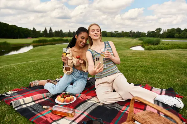 Two women enjoy a joyful picnic by a serene lake under fluffy clouds. — Stock Photo