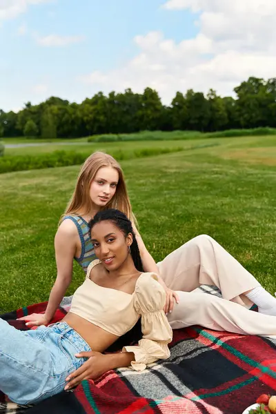 Two beautiful women relax together on a blanket, surrounded by green grass and blue skies. — Stock Photo