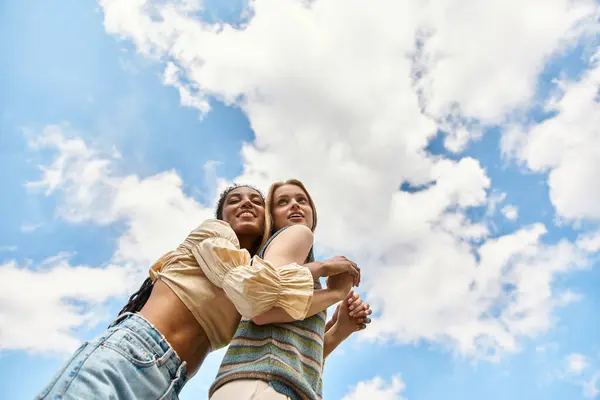 Two young women joyfully embrace outdoors under a sunny, cloudy sky. — Stock Photo