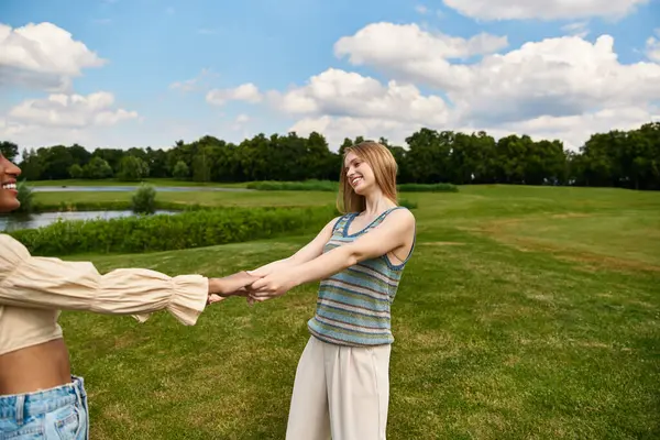 Two young women hold hands in a lush green park, radiating happiness under a bright sky. — Stock Photo