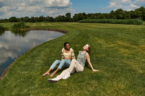 Two young women laugh and enjoy each others company by a peaceful lake under a blue sky — Stock Photo