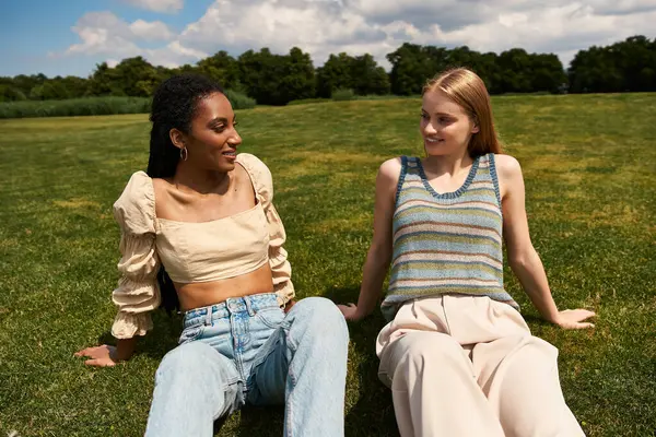 Two young women share laughter and joy on a grassy field under a bright blue sky, embracing love. — Stock Photo