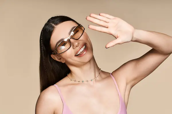 A cheerful young woman with a sun tan waves while wearing fashionable sunglasses, enjoying summer. — Stock Photo