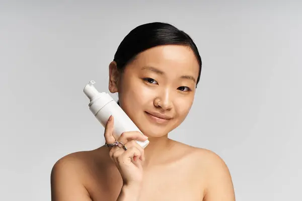 Young asian woman displays skincare product while smiling confidently in minimal studio backdrop. — Stock Photo