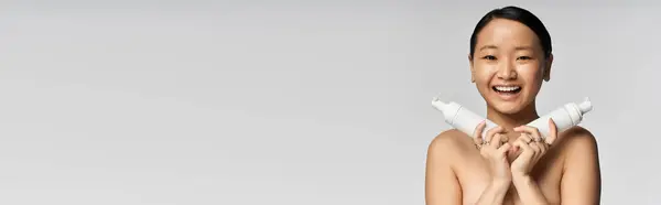 Radieuse jeune femme sourit tout en tenant des produits de soins de la peau, entourée d'un fond simple. — Photo de stock