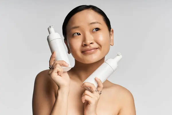 Una giovane donna asiatica sorride con gioia tenendo due prodotti per la cura della pelle su uno sfondo leggero. — Foto stock