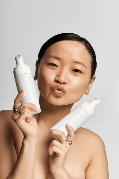 Smiling young asian woman poses playfully with skincare bottles in a bright studio setting. — Stock Photo
