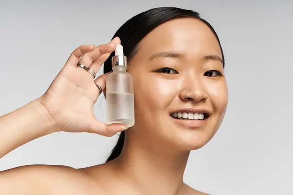 Young beautiful Asian woman smiles while showcasing a skincare serum against a soft background. — Stock Photo