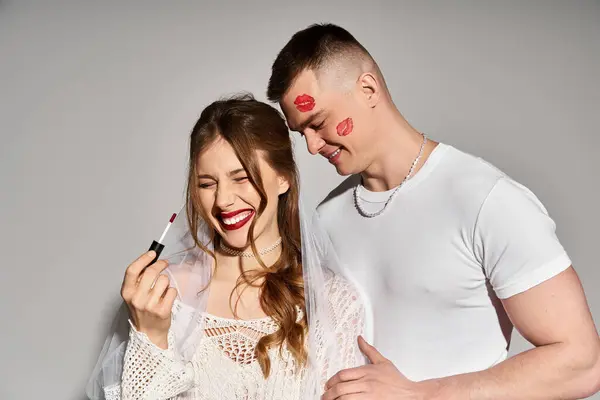 Una novia alegre sostiene el lápiz labial, sonriendo con su guapo novio a su lado. - foto de stock