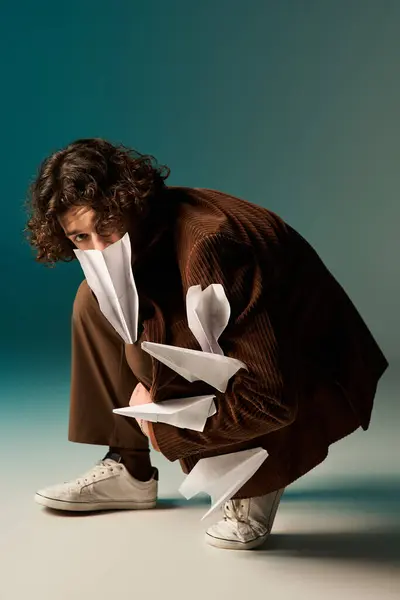 Young man in a brown corduroy blazer kneels down, playfully showcasing paper planes in studio. — Stock Photo