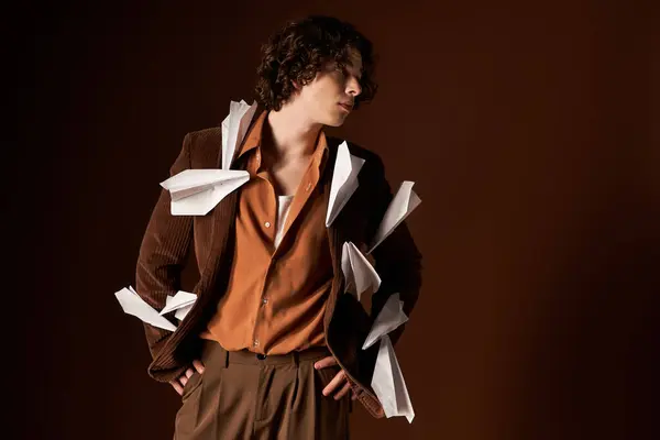 A handsome young man in a brown blazer poses with playful paper planes in a studio. — Stock Photo