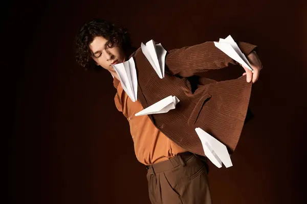 A young curly man in a brown corduroy blazer playfully displays paper planes, embodying creativity. — Stock Photo