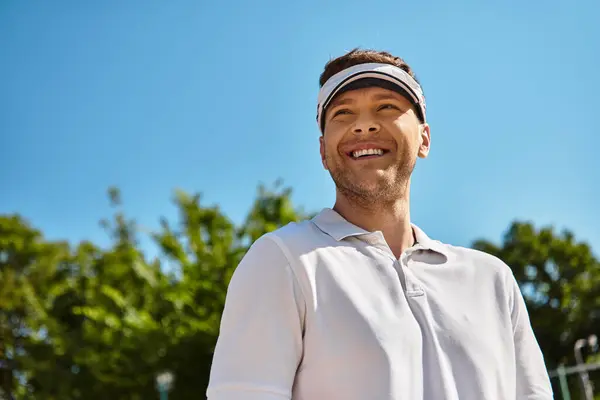 A fit man in white sportswear smiles brightly while playing tennis on a sunny day. — Stock Photo