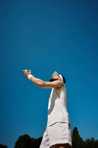 Un abile giocatore in abbigliamento sportivo bianco si prepara a servire una palla da tennis sotto un cielo blu chiaro. — Foto stock